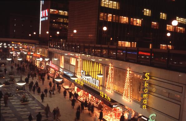 Kvällsvy med neonreklam över gågatan. I förgrunden bl a skobutikskedjan Skoman.